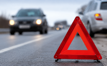 Ceļa malā novietota avārijas brīdinājuma zīme, fonā redzami pēc ceļu satiksmes negadījuma apstājušies automobiļi uz autoceļa.