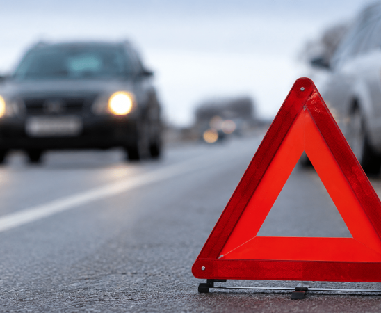 Ceļa malā novietota avārijas brīdinājuma zīme, fonā redzami pēc ceļu satiksmes negadījuma apstājušies automobiļi uz autoceļa.