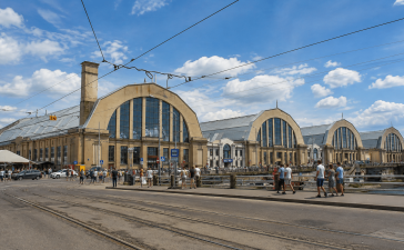 Rīgas Centrāltirgus Sakņu paviljons vasaras dienā ar cilvēkiem, tramvaja sliedēm un satiksmi pilsētas centrā.