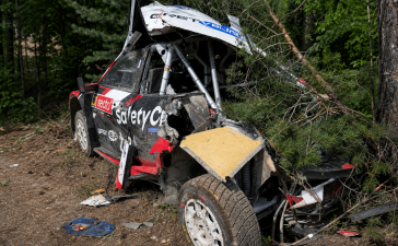 Smagi bojāta rallija drošības automašīna pēc avārijas meža posmā, redzami būtiski virsbūves bojājumi un nolauzti koki.